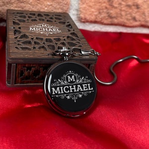 May include: A silver pocket watch with a black face and a chain. The watch face has an engraved design with the letter "M" and the name "MICHAEL".