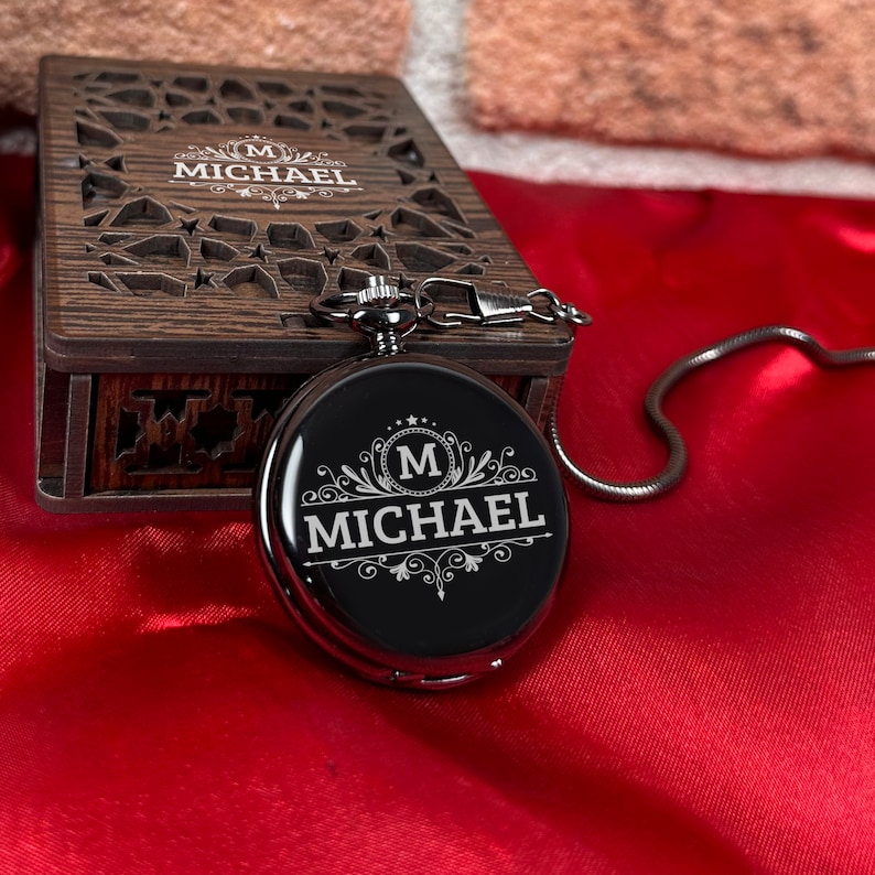 May include: A silver pocket watch with a black face and a chain. The watch face has an engraved design with the letter "M" and the name "MICHAEL".