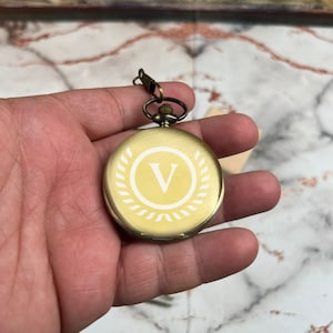 May include: A gold pocket watch with a white engraved letter V inside a wreath design.
