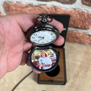 May include: A black pocket watch with a white clock face and a photo inside. The watch is open, revealing a color photograph. The watch is held in a hand, with a brick wall in the background.