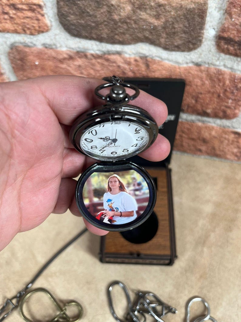 May include: A black pocket watch with a white clock face and a photo inside. The watch is open, revealing a color photograph. The watch is held in a hand, with a brick wall in the background.