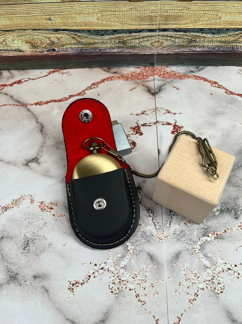 May include: A gold pocket watch with a black leather case and a red velvet lining. The watch is attached to a chain and a wooden block.