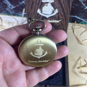 May include: A brass-colored pocket watch with an anchor and ship design, and the text "heavy cheddar". The watch is held in a hand, with a wooden box in the background featuring a similar anchor design.