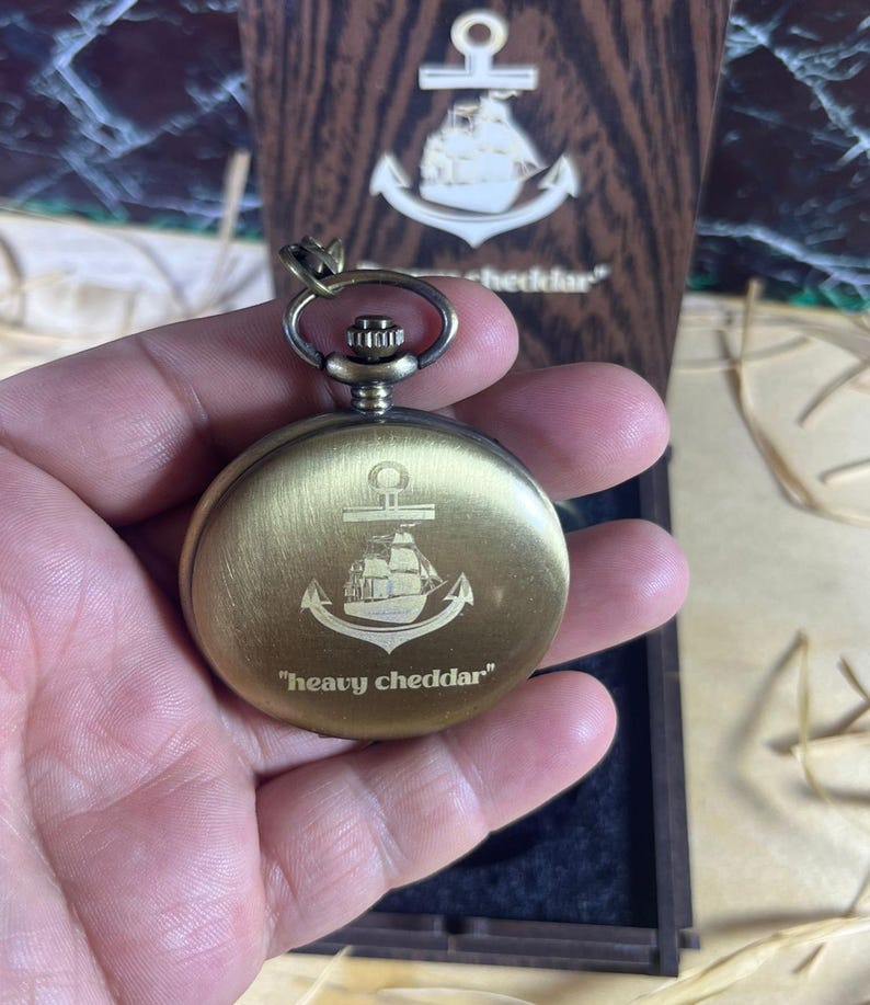 May include: A brass-colored pocket watch with an anchor and ship design, and the text "heavy cheddar". The watch is held in a hand, with a wooden box in the background featuring a similar anchor design.