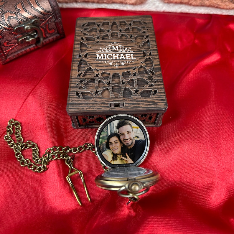 May include: A wooden box with a decorative cutout design and the name "Michael" engraved on the front. The box is open and a gold pocket watch with a photo of a couple inside is visible.