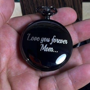 May include: A black pocket watch with the inscription "Love you forever Mom...". The watch has a circular face and a dark metal finish. The watch is held in a hand.