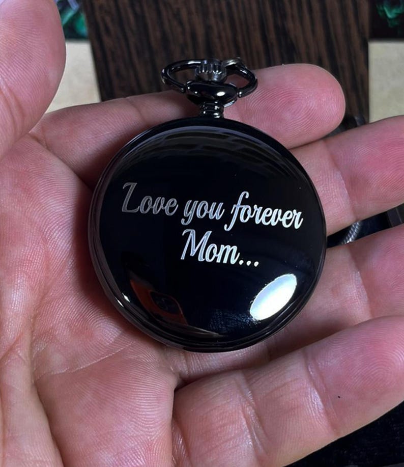 May include: A black pocket watch with the inscription "Love you forever Mom...". The watch has a circular face and a dark metal finish. The watch is held in a hand.