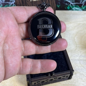 May include: A black pocket watch with the name "Brennan" engraved on the face. The watch has a dark metal finish and is held in a person's hand. The watch is round and has a decorative top. The watch is in a wooden box.