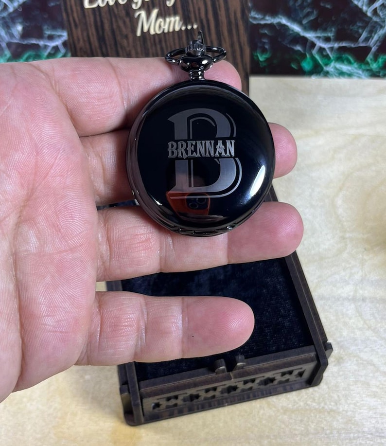 May include: A black pocket watch with the name "Brennan" engraved on the face. The watch has a dark metal finish and is held in a person's hand. The watch is round and has a decorative top. The watch is in a wooden box.