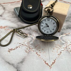 May include: A vintage-style pocket watch with a white face and black Roman numeral hour markers. The watch has a gold-tone finish and is attached to a chain. The watch is sitting on a wooden surface.