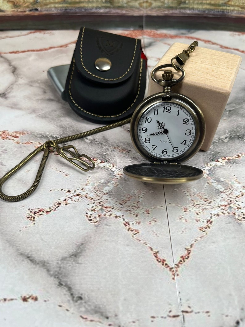 May include: A vintage-style pocket watch with a white face and black Roman numeral hour markers. The watch has a gold-tone finish and is attached to a chain. The watch is sitting on a wooden surface.