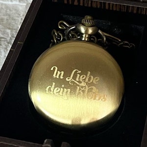May include: A gold pocket watch with a chain in a black velvet box. The watch face has the inscription "In Liebe dein Boss" engraved on it.