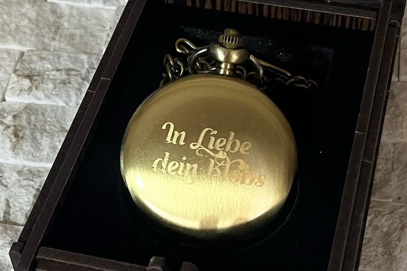 May include: A gold pocket watch with a chain in a black velvet box. The watch face has the inscription "In Liebe dein Boss" engraved on it.