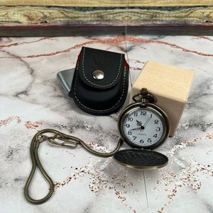 May include: A gold pocket watch with a white face and black Roman numeral hour markers. The watch is on a gold chain and has a black leather case with a gold button.