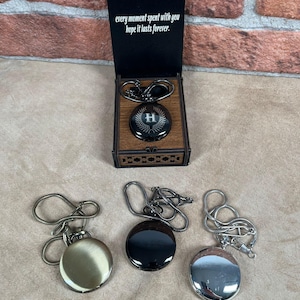 May include: A collection of pocket watches in various finishes: gold, black, and silver. One watch is displayed in a wooden box with the text "every moment spent with you hope it lasts forever." The watch in the box has an "H" initial.