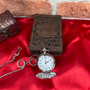 May include: A silver pocket watch with a white face and black numbers. The watch is open and the chain is attached to a small, wooden box with a carved design.