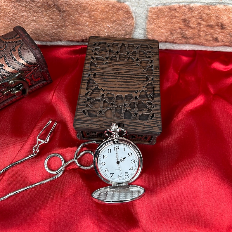 May include: A silver pocket watch with a white face and black numbers. The watch is open and the chain is attached to a small, wooden box with a carved design.