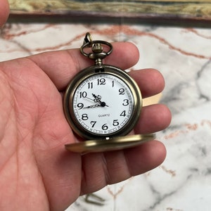 May include: A gold-toned pocket watch with a white face and black Roman numeral hour markers. The watch has a quartz movement and a small, round, gold-toned case.
