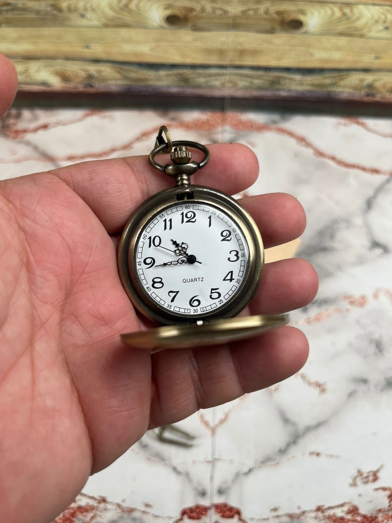 May include: A gold-toned pocket watch with a white face and black Roman numeral hour markers. The watch has a quartz movement and a small, round, gold-toned case.