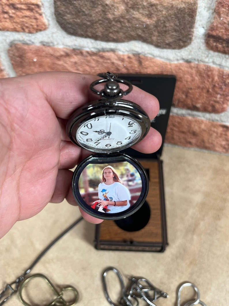 May include: A black pocket watch with a white clock face and a photo inside. The watch is open, revealing a color photograph. The watch is held in a hand, with a brick wall in the background.