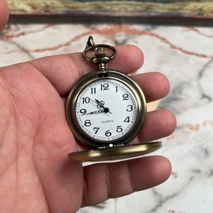 May include: A gold-toned pocket watch with a white face and black hands. The watch has a round case and a chain attached to the top. The watch face has black Roman numeral hour markers and black minute markers. The watch face also has the word "QUARTZ" printed on it.