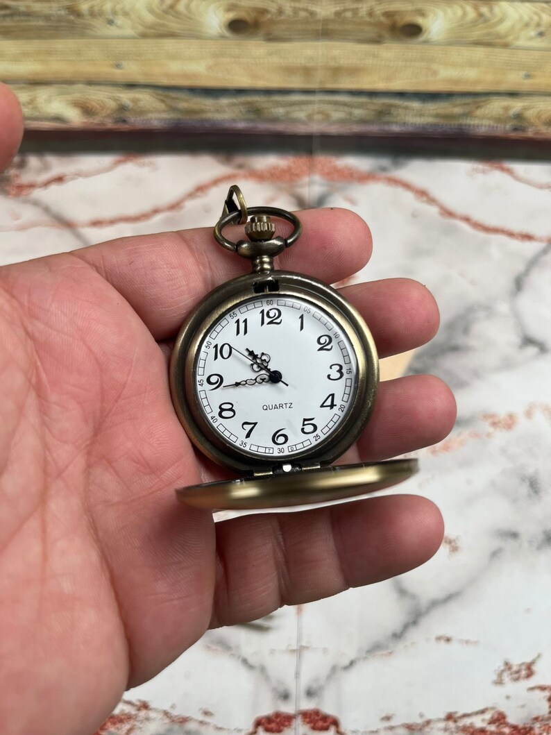 May include: A gold-toned pocket watch with a white face and black hands. The watch has a round case and a chain attached to the top. The watch face has black Roman numeral hour markers and black minute markers. The watch face also has the word "QUARTZ" printed on it.