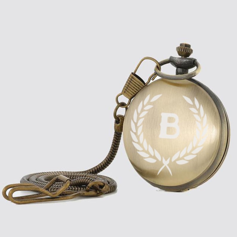 May include: A gold-toned pocket watch with a chain. The watch face has a white laurel wreath design with the letter "B" in the center.
