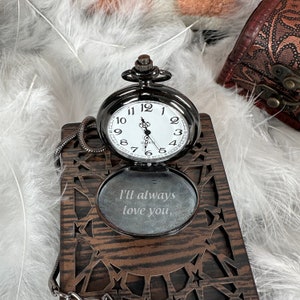 May include: A black pocket watch with a white face and black hands. The watch is open and the inside of the case is engraved with the words "I'll always love you".