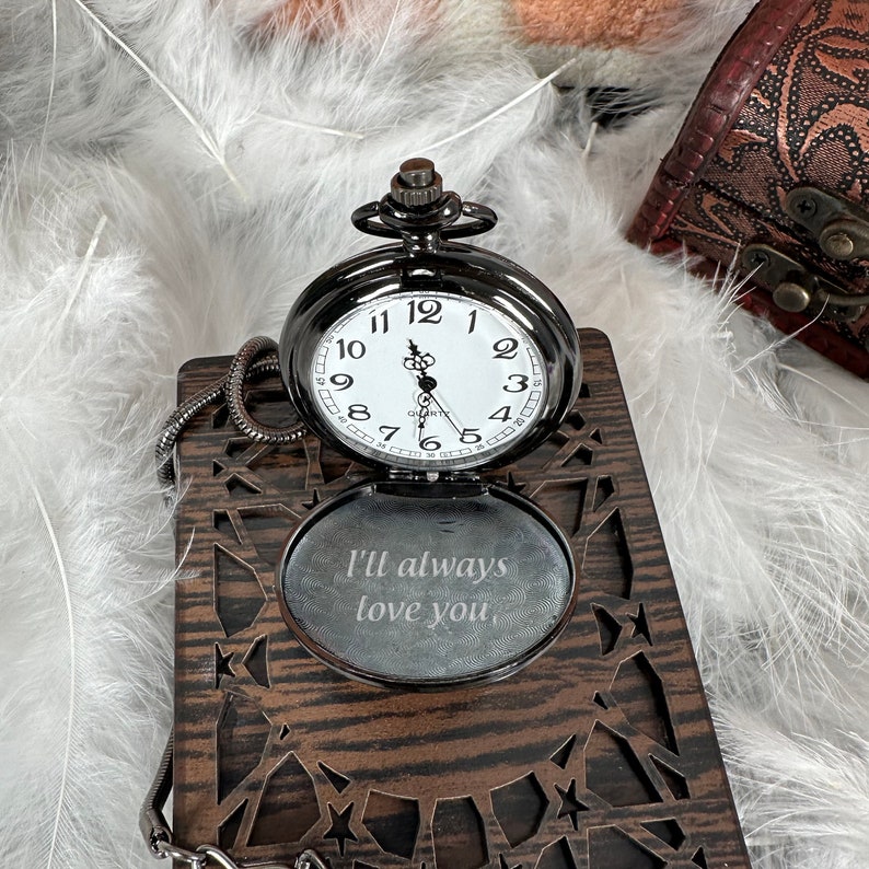 May include: A black pocket watch with a white face and black hands. The watch is open and the inside of the case is engraved with the words "I'll always love you".