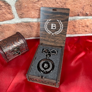 May include: A black pocket watch with a silver engraved letter B inside a laurel wreath design. The watch is in a wooden box with a similar engraved design on the lid. The box is on a red surface.