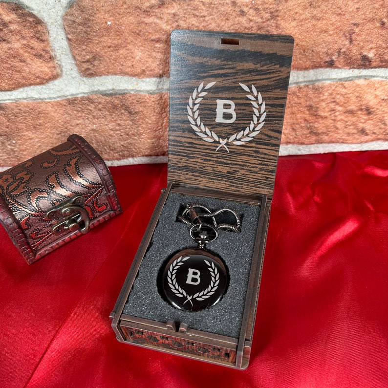 May include: A black pocket watch with a silver engraved letter B inside a laurel wreath design. The watch is in a wooden box with a similar engraved design on the lid. The box is on a red surface.