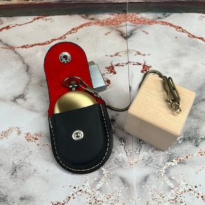 May include: A black leather pocket watch case with a gold pocket watch inside. The case has a red lining and a gold snap closure. The watch is attached to a gold chain with a wooden block at the end.