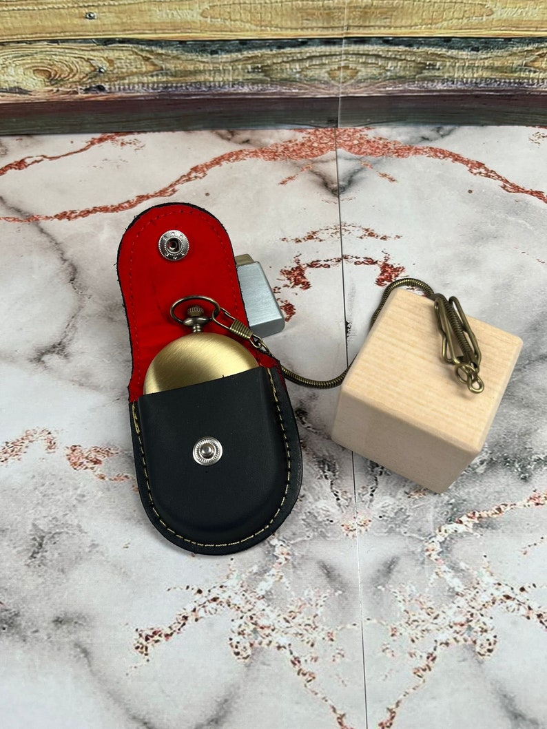 May include: A black leather pocket watch case with a gold pocket watch inside. The case has a red lining and a gold snap closure. The watch is attached to a gold chain with a wooden block at the end.