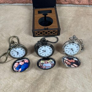 May include: Three pocket watches with oval photo inserts. One watch is bronze, one is black, and one is silver. The watch faces have black numbers on a white background. A wooden box with a watch stand is in the background.