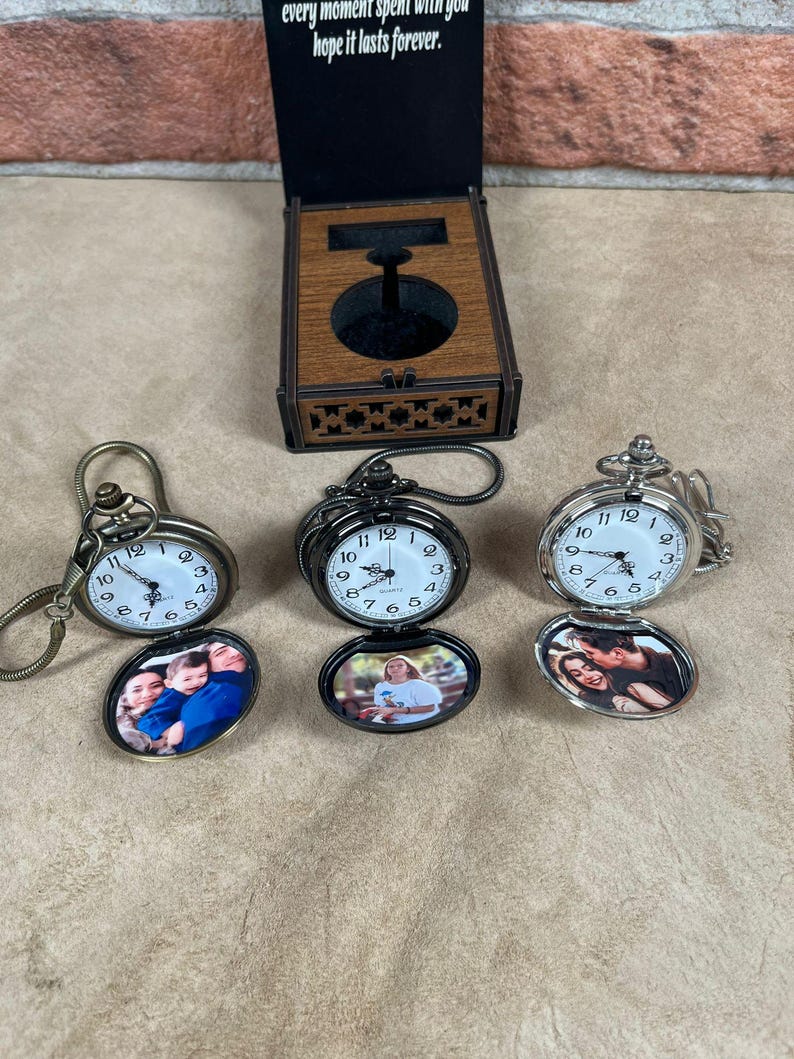 May include: Three pocket watches with oval photo inserts. One watch is bronze, one is black, and one is silver. The watch faces have black numbers on a white background. A wooden box with a watch stand is in the background.
