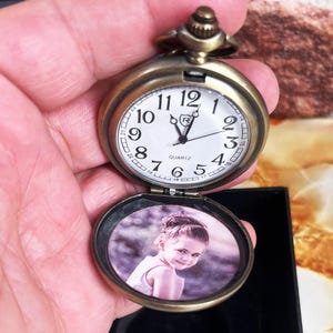 May include: Antique bronze-colored pocket watch with a white clock face and black numerals. The watch is open, revealing a small photo of a child. The watch has a quartz movement and a decorative crown.