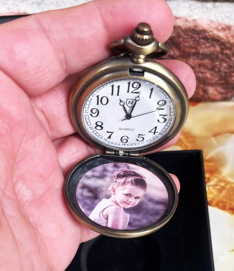 May include: Antique bronze-colored pocket watch with a white clock face and black numerals. The watch is open, revealing a small photo of a child. The watch has a quartz movement and a decorative crown.