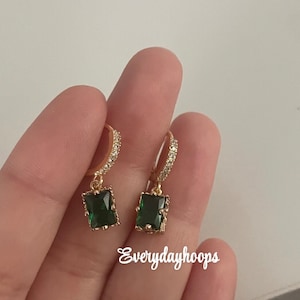 May include: Gold hoop earrings with a dangling emerald green square stone. The earrings are adorned with small clear stones.
