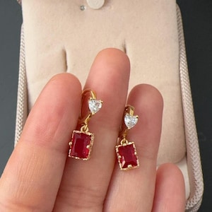 以下が含まれることがあります： 赤と透明なキュービックジルコニアの石が付いた、金色のイヤリングのペア。イヤリングはハート型のトップに、長方形の石がぶら下がっています。