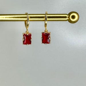 May include: A pair of gold-colored hoop earrings, each featuring a rectangular, deep red gemstone. The gemstones are set within a gold-colored frame. The earrings are displayed on a gold-colored bar against a light background.