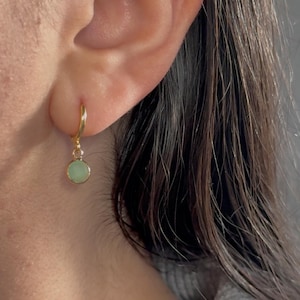 May include: A gold-coloured hoop earring with a small, round, pale green charm. The earring is attached to an ear. The charm is a light, pastel green colour. The earring is a simple, elegant design.