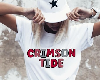 May include: A white t-shirt with the words "CRIMSON TIDE" in red and grey block letters. The person is wearing a white baseball cap with black star accents and blue jeans. The background is a textured, off-white wall.