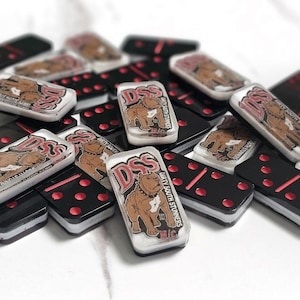 Könnte beinhalten: Eine Ansammlung von Dominosteinen mit schwarzen Basen und roten Punkten. Einige Dominosteine haben eine transparente Oberseite mit einer braunen Hunde-Grafik und dem Text "DSS Dirty South Stunners".