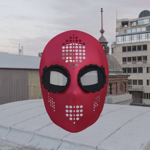 Op de afbeelding: Een rood masker met zwarte oog- en monddetails en een patroon van kleine gaatjes. Het masker staat tegen een stadslandschap achtergrond, met gebouwen en een bewolkte lucht.