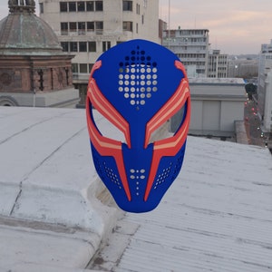 Pode incluir: Uma máscara azul e vermelha com um design geométrico. A máscara tem pequenos orifícios de ventilação e aberturas para os olhos. A máscara é feita de um material duro, provavelmente para cosplay ou fantasia.