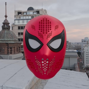 May include: A bright red full-face mask featuring a perforated pattern across the forehead and chin area. The mask includes two large, almond-shaped eye openings outlined in thick black frames with a white mesh screen covering the eye area.