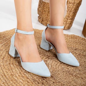 以下が含まれることがあります： 足首ストラップ付きの薄いブルーのスエードのヒールサンダル。サンダルはブロックヒールとポインテッドトゥです。