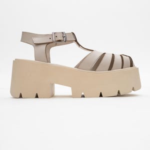 Könnte beinhalten: Beige Plateausandalen mit T-Riemen-Design und Schnallenverschluss. Die Sandalen haben eine dicke, klobige Sohle und eine neutrale Farbpalette, die für verschiedene Outfits geeignet ist. Die Sandalen sind aus glattem Material.