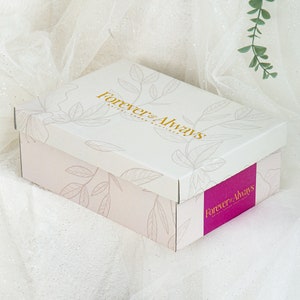 Op de afbeelding: Een witte cadeaudoos met een roze bloemenpatroon en de tekst "Forever & Always Bridal Boutique" in gouden letters. De doos is gesloten en staat op een wit oppervlak.