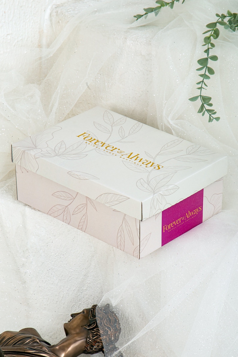 Op de afbeelding: Een witte cadeaudoos met een roze bloemenpatroon en de tekst "Forever & Always Bridal Boutique" in gouden letters. De doos is gesloten en staat op een wit oppervlak.